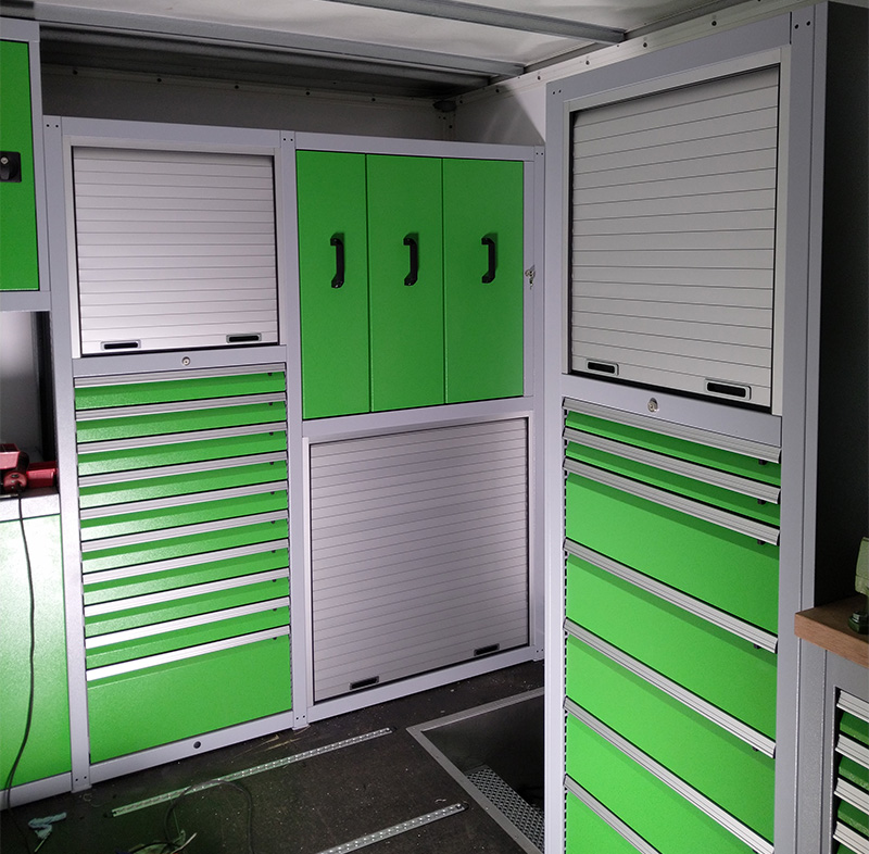 Innenansicht einer modularen Werkstatteinrichtung mit mehreren grünen Schrankmodulen, Schubladenschränken und silbernen Rollladenklappen.
